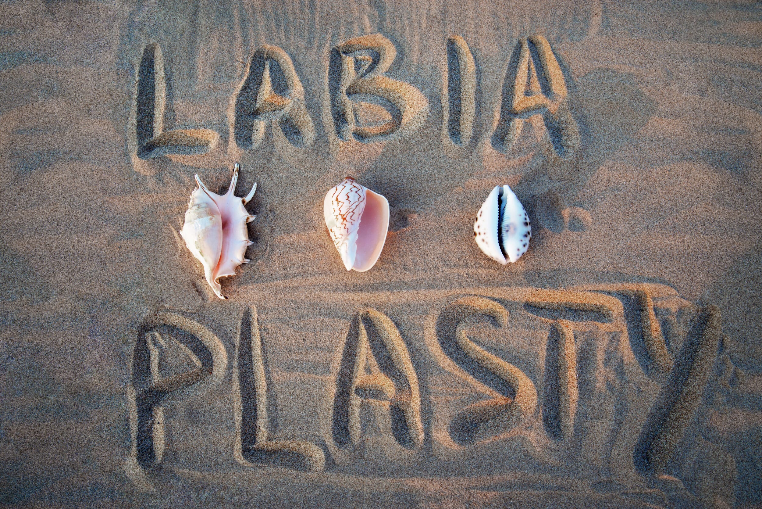 The word “Labiaplasty” written in the sand with 3 seashells lined up next to it